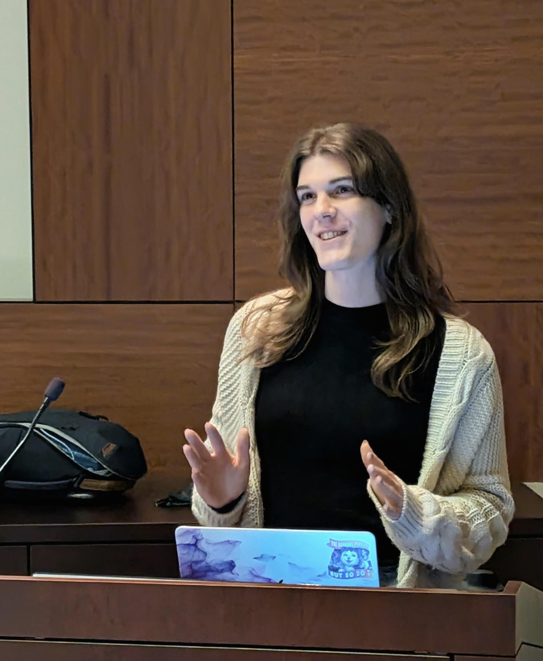 An image of Nora Mahon speaking at a podium. She is a young woman with brown hair, wearing a cardigan and black shirt.